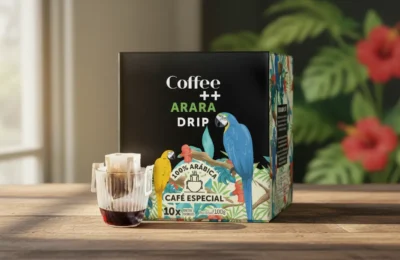 Caixa de café Arara Drip ao lado de copo de vidro com sachê de café coando sobre mesa de madeira.