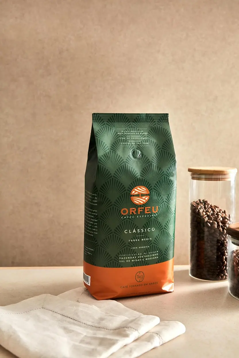Grãos de café Orfeu Clássico em close-up, espalhados em uma superfície branca, com a embalagem ao fundo.